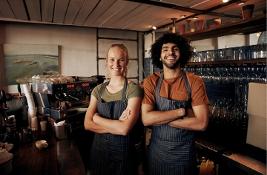 Portrait of young female and male staff wearing apron standing behind counter in cafe with crossed hands looking at camera